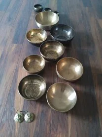 Une douzaine de bols tibétains alignés sur un parquet, avec des cymbalettes au premier plan